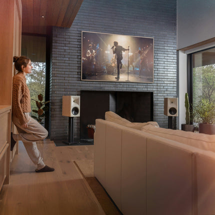 Pereche Klipsch The Nines II in finisaj Oak, pe stative, langa un televizor care reda un concert